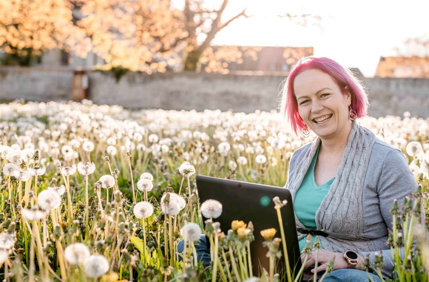 Katja Gersdorf sitzt lächelnd mit dem Laptop in einer Pusteblumenwiese